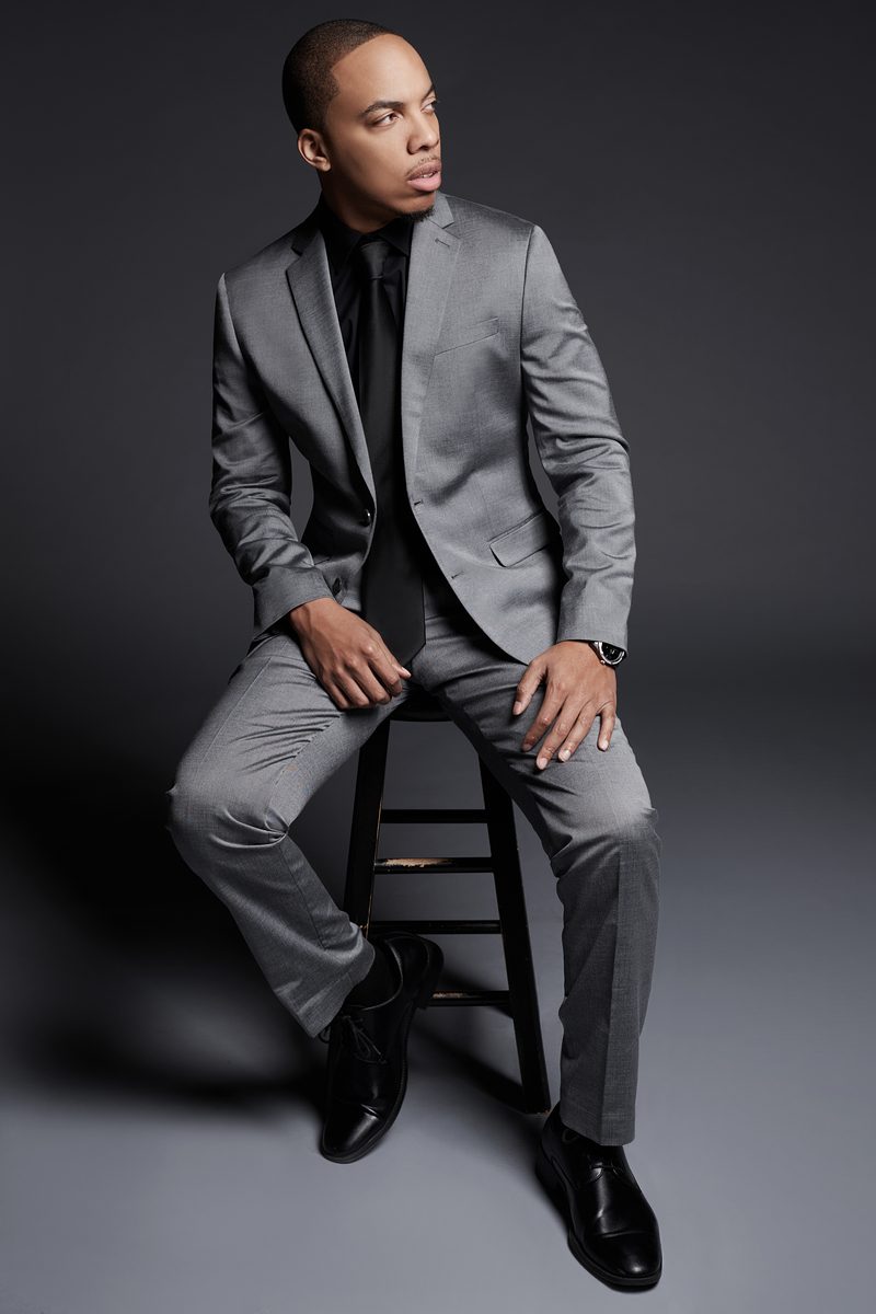 Full-length studio portrait of Jerel Crockett seated on a stool in a grey suit and black shirt against a grey seamless backdrop.