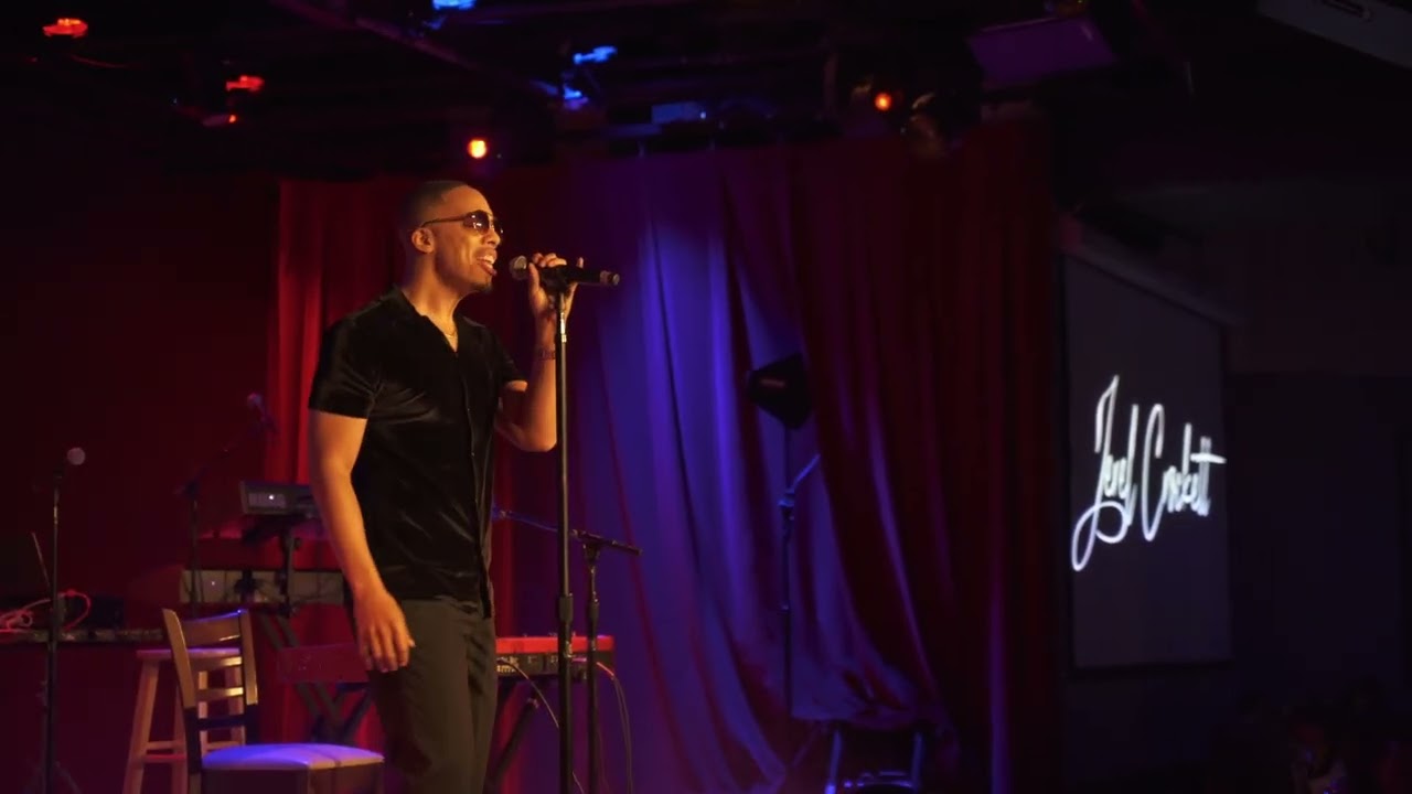 Jerel Crockett front of stage at City Winery, Washington, D.C., singing into a handheld microphone under red and blue light, his script logo projected on the screen behind him.